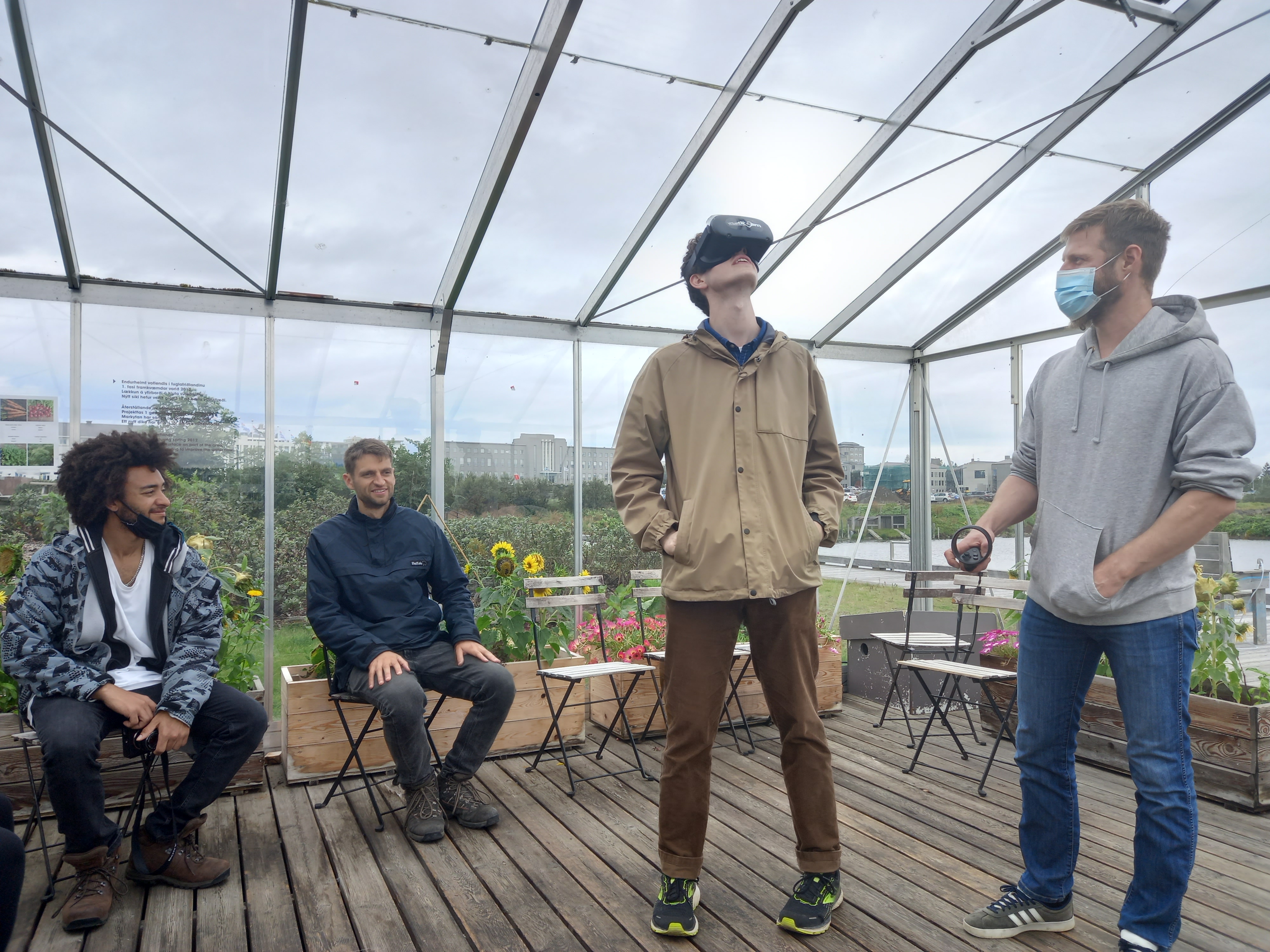 Strákur stendur inni í gróðurhúsi með sýndarveruleikagleraugu á höfðinu og horfir upp í loft. Tveir strákar sitja fyrir aftan hann á stólum. Piotr Loj stendur hjá honum.